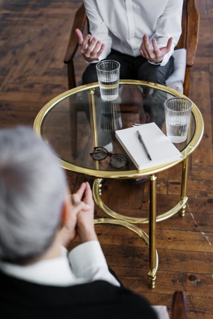 woman-in-black-long-sleeve-shirt-sitting-on-brown-wooden-chair-4098369 Two adults discussing mental health in a counseling session across a glass table indoors.